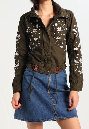 Femme porte une veste vert foncé brodée de fleurs avec des cordons et une jupe en jean bleue avec fermeture éclair devant.