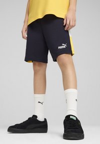Marinblå shorts med gula och vita detaljer, prydda med en logotyp på låret. Svarta sneakers med textilstruktur och vita strumpor.
