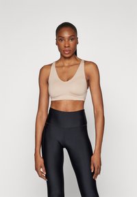 Beige sportsbh med V-hals, brede stropper og sømløst design. Kombinert med høydeplassering svarte leggings som har en glatt tekstur.
