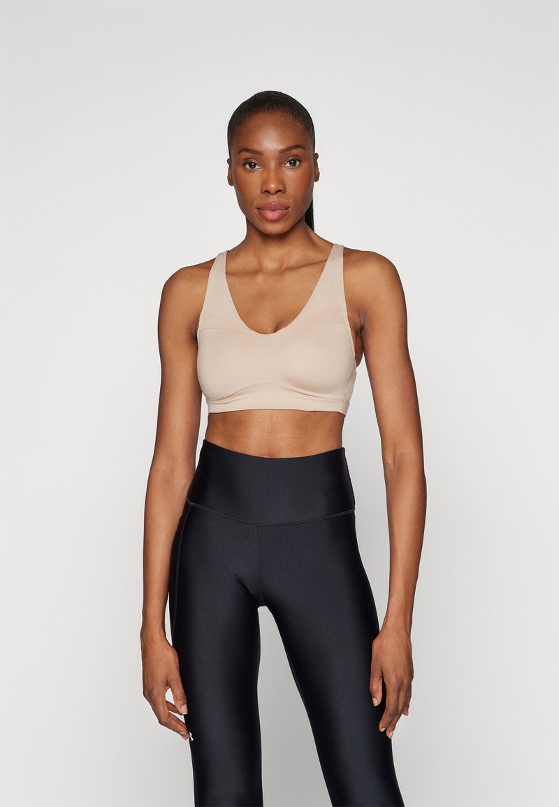 Beige sportsbh med V-hals, brede stropper og sømløst design. Kombinert med høydeplassering svarte leggings som har en glatt tekstur.