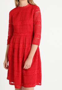 Robe en dentelle rouge avec un col rond, des manches trois quarts et une jupe évasée. Présente des motifs floraux complexes et une surface texturée.