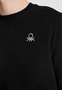 Svart tröja med ribbad rund halsringning och en liten grå broderad logotyp på bröstet. Tyget ser slätt och mjukt ut.