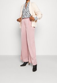 Pantaloni a gamba larga rosa chiaro abbinati a una blusa floreale e a un cardigan crema, accentuati da stivali con tacco neri. Texture morbide e vestibilità sartoriale.