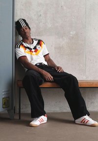 Νεαρός άνδρας φοράει φανέλα ποδοσφαίρου της Γερμανίας, μαύρο παντελόνι, σκούφο και λευκά sneakers, κάθεται χαλαρά σε μια ξύλινη παγκίδα δίπλα σε σκυρόδεμα τοίχο.