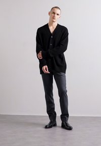 Cárdigan negro de punto con escote en V, con tres botones y bolsillos. Combinado con pantalones gris oscuro y botas de cuero negras.