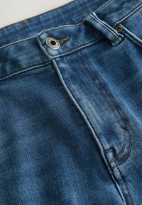 Farkut sinisellä pesulla, varustettuna vetoketjulla, metallinappikiinnityksellä ja näkyvillä ompeluosoilla vyötärön ja taskujen reunoilla.