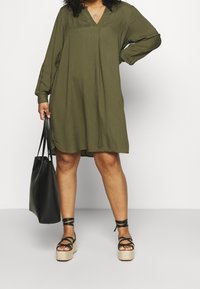 Olivengrünes langärmeliges Kleid mit V-Ausschnitt, das einen lockeren Schnitt hat. Getragen mit schwarzen, schnürbaren Espadrilles und einer schwarzen Tragetasche.