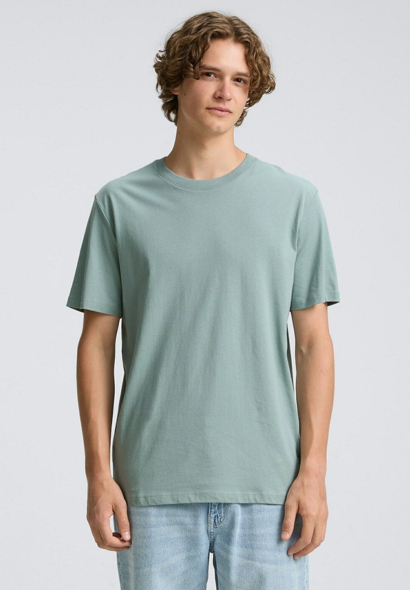 Jasnozielona bawełniana koszulka z krótkim rękawem i okrągłym dekoltem. Prosty design o luźnym kroju. Noszona z jasnoniebieskimi dżinsami.