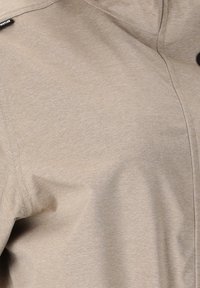 Nahaufnahme eines beigen Stoffs mit sichtbaren Nähten und Detail der Nahtarbeiten, zeigt ein kleines schwarzes Etikett und einen teilweisen schwarzen Knopf am Rand.