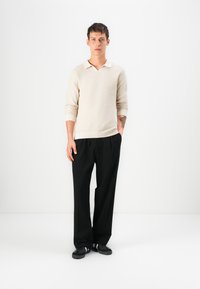 Beige strikket genser med V-hals og lange ermer, kombinert med sorte plissert bukser og sorte joggesko med hvite striper.