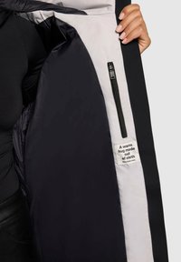 Zwarte geïsoleerde jas met een gladde textuur, voorzien van een lichtgrijze voering en een ritszak, gelabeld "Een warme omhelzing van stof."