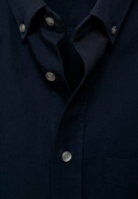 Navy button-down shirt met een puntige kraag, zes zilveren knopen en een borstzak. De stof lijkt soepel en gestructureerd.