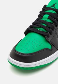 Gros plan sur l'avant d'une basket verte et noire avec des perforations, des lacets noirs, une semelle intermédiaire blanche et une semelle extérieure verte sur fond blanc.