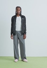 Zwarte jas met patroon en witte accenten, witte tanktop, grijze broek en roze schoenen. Model heeft gevlochten haar en staat op groene vloer.