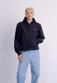 Black hooded sweatshirt with a front pocket and logo detail, paired with light blue pants. Smooth fabric with a relaxed fit design.