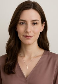 Silverhalsband med en avlång berlock, bärs över en mauve V-ringad blus. Modellen har mellanbrunt hår och minimal sminkning.