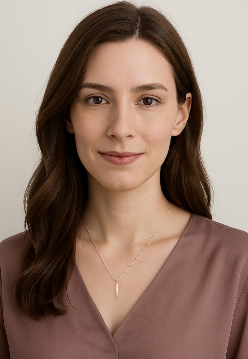 Silverhalsband med en avlång berlock, bärs över en mauve V-ringad blus. Modellen har mellanbrunt hår och minimal sminkning.