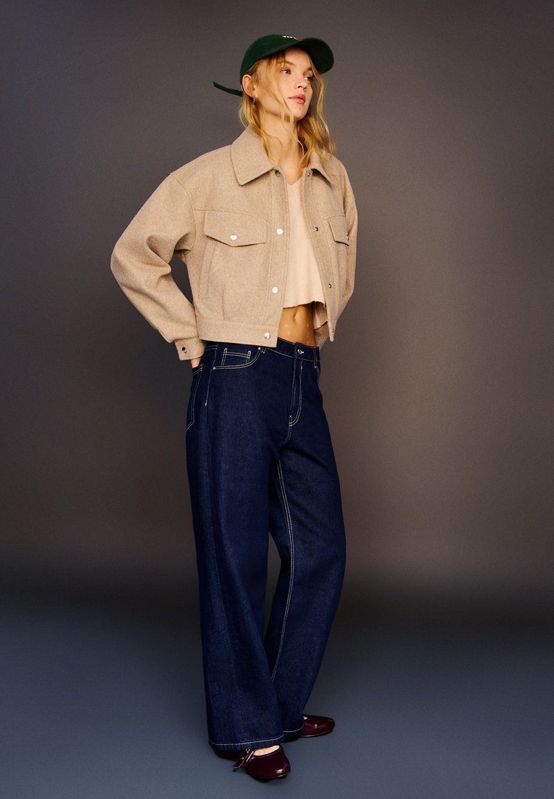 Beige cropped jacket with chest pockets, light knit top, wide-leg dark denim jeans, and maroon shoes; green cap completes the look.