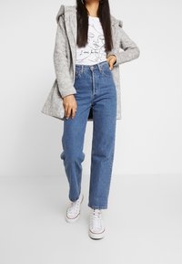 Femme portant un manteau gris à capuche, un t-shirt blanc à motifs, un jean bleu taille haute et des baskets blanches, debout avec une main sur la hanche.