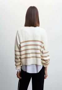Cremfarbener und beiger gestreifter Pullover mit gerippter Textur, verkürztem Design, weiten Ärmeln und rundem Ausschnitt, über einem weißen Hemd getragen.