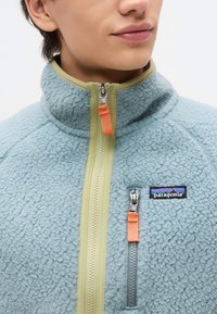 Κοντινό πλάνο ενός ατόμου που φοράει ανοιχτό μπλε φλις μπουφάν με μπεζ φερμουάρ και πορτοκαλί τσεπάκια φερμουάρ, με το λογότυπο της Patagonia στην τσέπη στο στήθος.