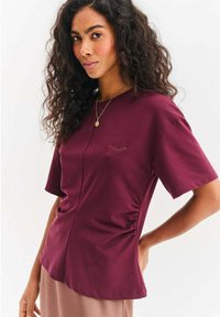 Bordowy top z krótkim rękawem wykonany z miękkiego materiału, z okrągłym dekoltem. Posiada marszczenia po bokach oraz mały haftowany detal przypominający rzęsy.