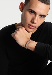 Sølv metallarmbånd båret på håndleddet, med en jevn, polert overflate. Modellen har kort hår og er kledd i en svart langermet topp.