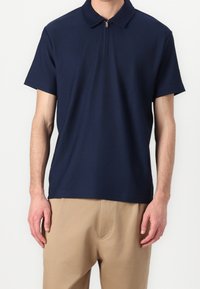 Marinblå ribbad polo skjorta med dragkedja i kragen, korta ärmar och en mjuk textur. Kombinerad med ljusbeiga byxor.