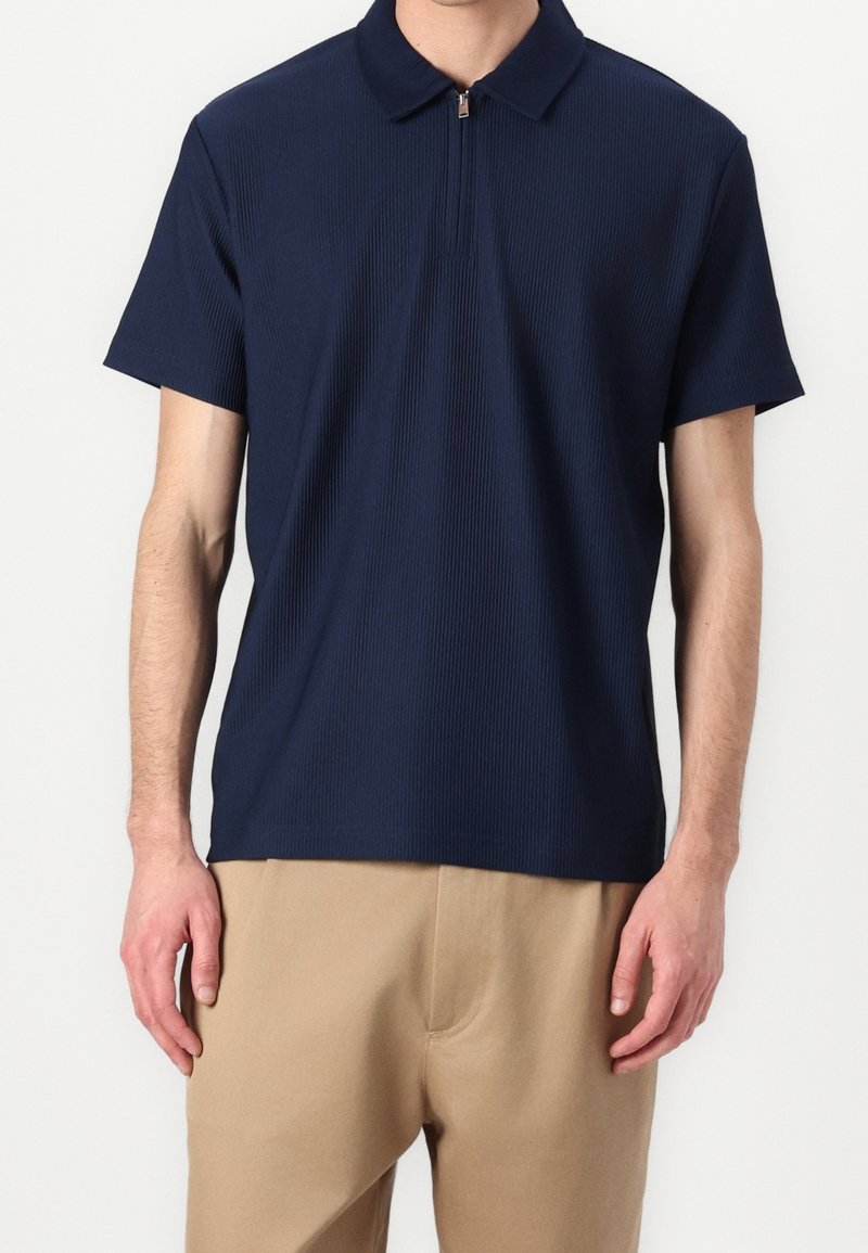 Marinblå ribbad polo skjorta med dragkedja i kragen, korta ärmar och en mjuk textur. Kombinerad med ljusbeiga byxor.