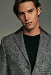 Grå tekstureret blazer med en brun corduroy-krave, der har en enkelt knaplukning og en lomme på venstre bryst, båret over en mørkegrøn sweater.