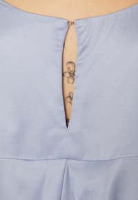 Ljubičasta bluza s V-izrezom na leđima, koja ima mali metalni kopča. Glatka tkanina s suptilnom teksturom i detaljima šivanja.