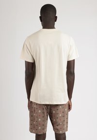 Camiseta de algodón color beige claro con mangas cortas, combinada con pantalones cortos marrones que presentan un patrón repetido de varios pequeños diseños.