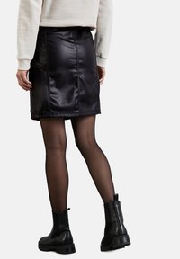 Černá sukně z umělé kůže s přiléhavým střihem, vertikálními švy a rozparkem na zadní straně, spárovaná s průhlednými černými punčochami a kotníkovými botami.