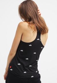 Black tank top made of cotton, featuring a repeated shoe graphic pattern in white. Racerback design with a small tag detail at the hem.