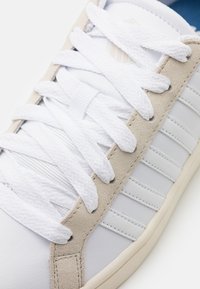 White sneaker with a smooth leather upper and beige suede accents. Features white flat laces, a textured tongue, and a rubber sole.
