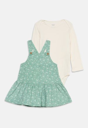 Body bébé crème à manches longues associé à une robe tablier verte à motif floral avec bretelles boutonnées.
