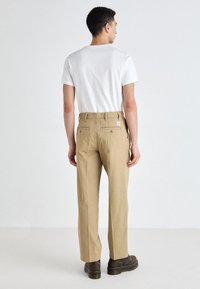 Beige cotton trousers with a straight cut, two back pockets, and a small logo tag, paired with a plain white t-shirt and dark shoes.