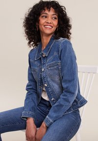 Veste en denim bleu moyen, avec un design classique comprenant des poches avant et des boutons en métal argenté. Associée à un jean bleu assorti.