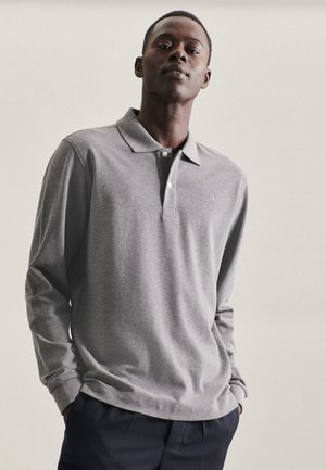 Grijze polo met lange mouwen, kraag, knoopsluiting en geborduurd logo. De stof lijkt zacht en textuurrijk en is gecombineerd met donkere broeken.