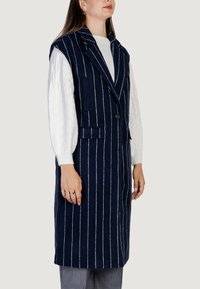 Námořnická pruhovaná vesta s klopovým límcem, zapínáním na knoflíky a dvěma předními kapsami. Nositá přes bílý svetr s kabelovým pletením.