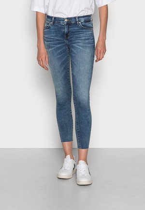 Jean skinny en denim bleu moyen, avec une taille haute, un délavage subtil et une texture lisse, associé à des baskets blanches.