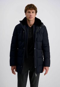 Man in een zwarte gewatteerde winterjas over een zwart shirt en grijze broek, staande tegen een effen witte achtergrond.