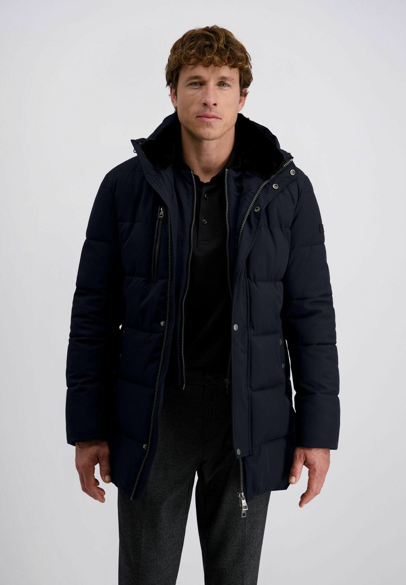 Man in een zwarte gewatteerde winterjas over een zwart shirt en grijze broek, staande tegen een effen witte achtergrond.