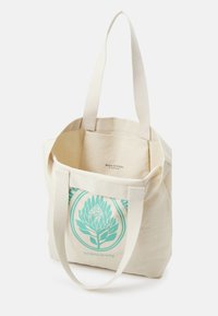 Borsa tote in tela beige con un design botanico in turchese chiaro e testo, dotata di due manici robusti e apertura superiore. Nome del marchio visibile all'interno.