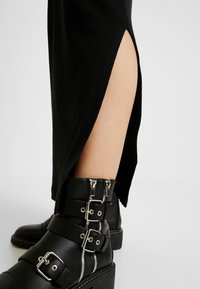 Jupe noire côtelée avec une fente latérale, associée à des bottines noires avec boucles et zips. Accent sur les textures et les matériaux contrastés.