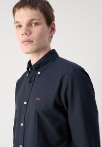 Marineblauw overhemd met knopen, gemaakt van een stevige stof met witte knopen en een klein rood "BOSS" logo op de borst. Voorzien van een standaardkraag en lange mouwen.