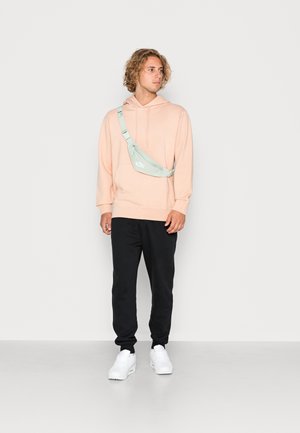 Felpa rosa chiaro, jogger neri, sneakers bianche; il modello indossa una borsa a tracolla verde menta con logo bianco. Tessuto morbido e vestibilità rilassata.
