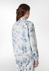 Jachetă cu model floral într-o nuanță de albastru deschis, confecționată dintr-un material moale, cu mâneci lungi, manșete elastice și un etichetă roz la gulerul din spate.