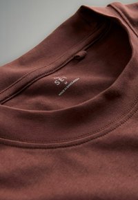 Camiseta de algodón marrón con cuello redondo, detalles de costura y una etiqueta que dice "SEI", con la inscripción "Hecho en Bangladesh".