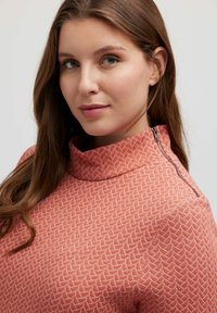 Maglione in tessuto coral con colletto alto e dettaglio con zip. Presenta un motivo geometrico bianco. Tessuto morbido con una leggera lucentezza.